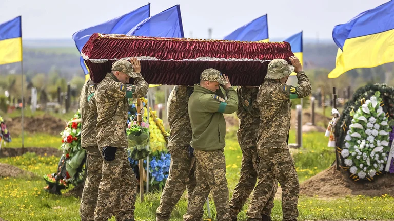 Feretro cargado por soldados ucranianos en uniforme de combate entre banderas y flores.
